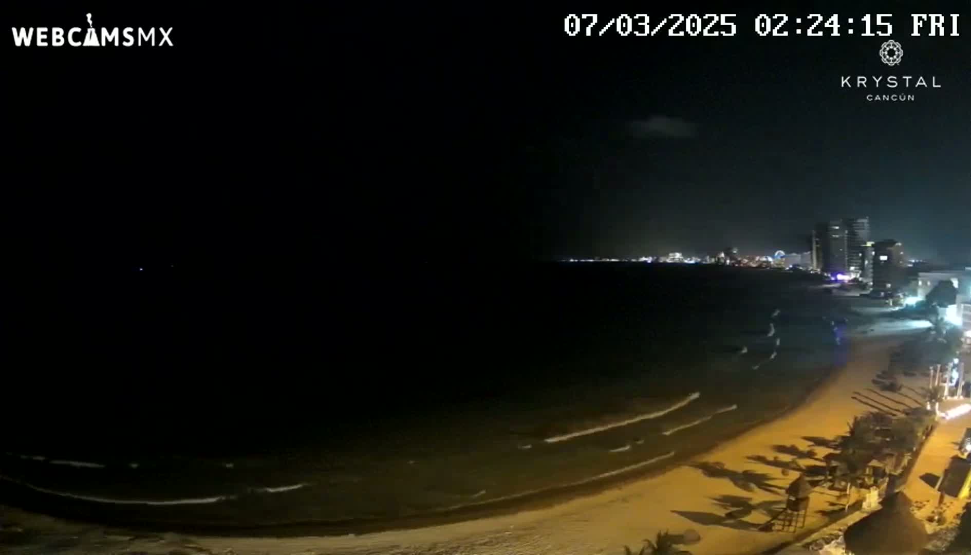 A nighttime beach scene shows dark water with small waves lapping a sandy shore, with city lights and buildings visible in the distance under a mostly clear sky.