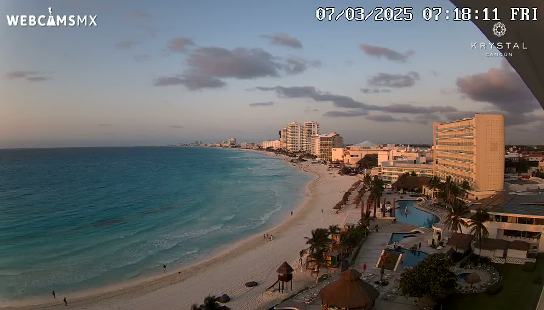 A mostly clear sunset sky overlooks a beach with turquoise water, hotels, and palm trees.