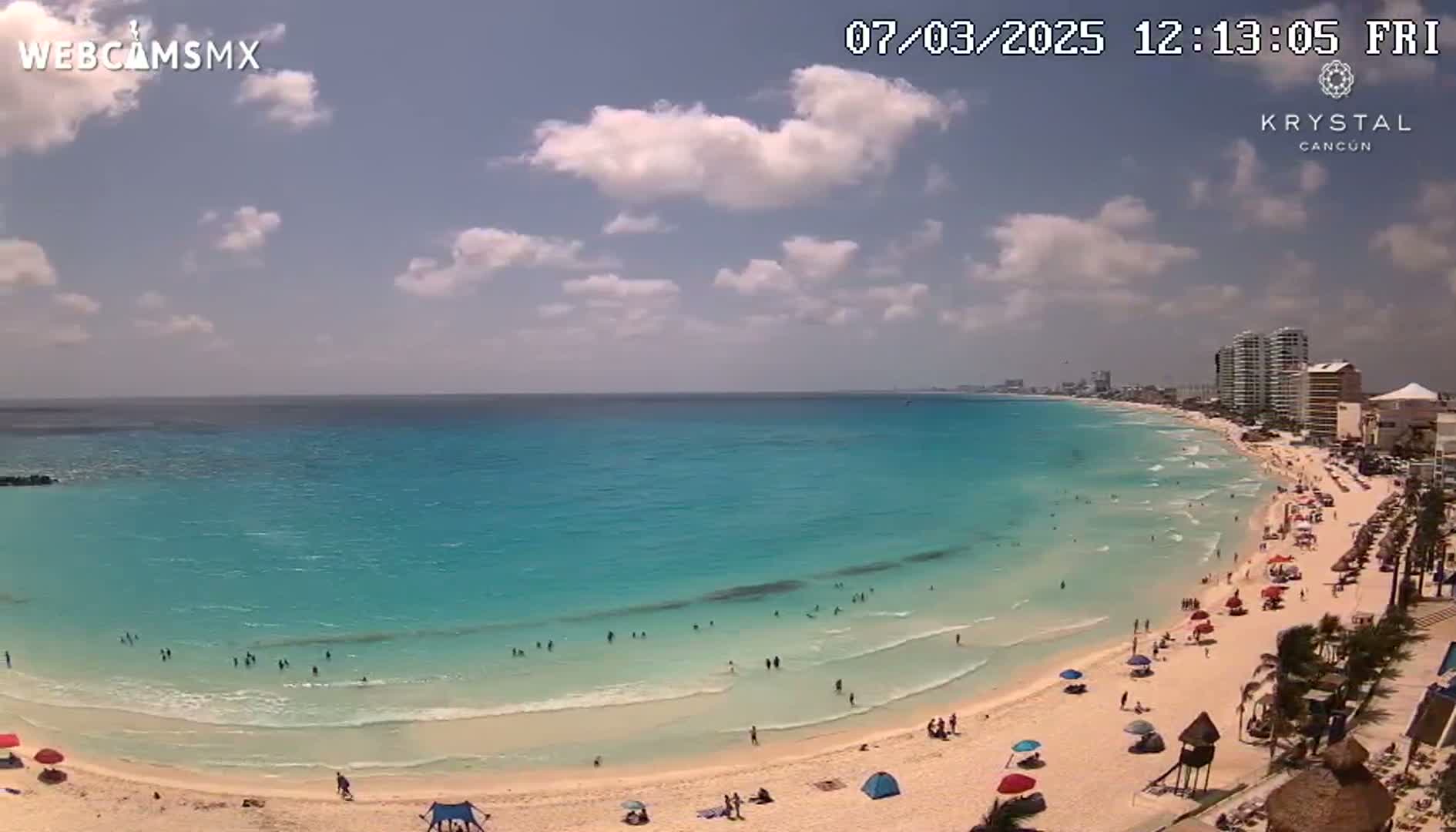 A partly cloudy sky overlooks a beach with turquoise water and many people swimming and relaxing under umbrellas.