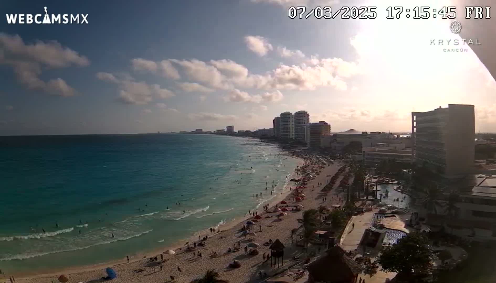 A partly cloudy sunset illuminates a beach with many people, hotels, and turquoise water.