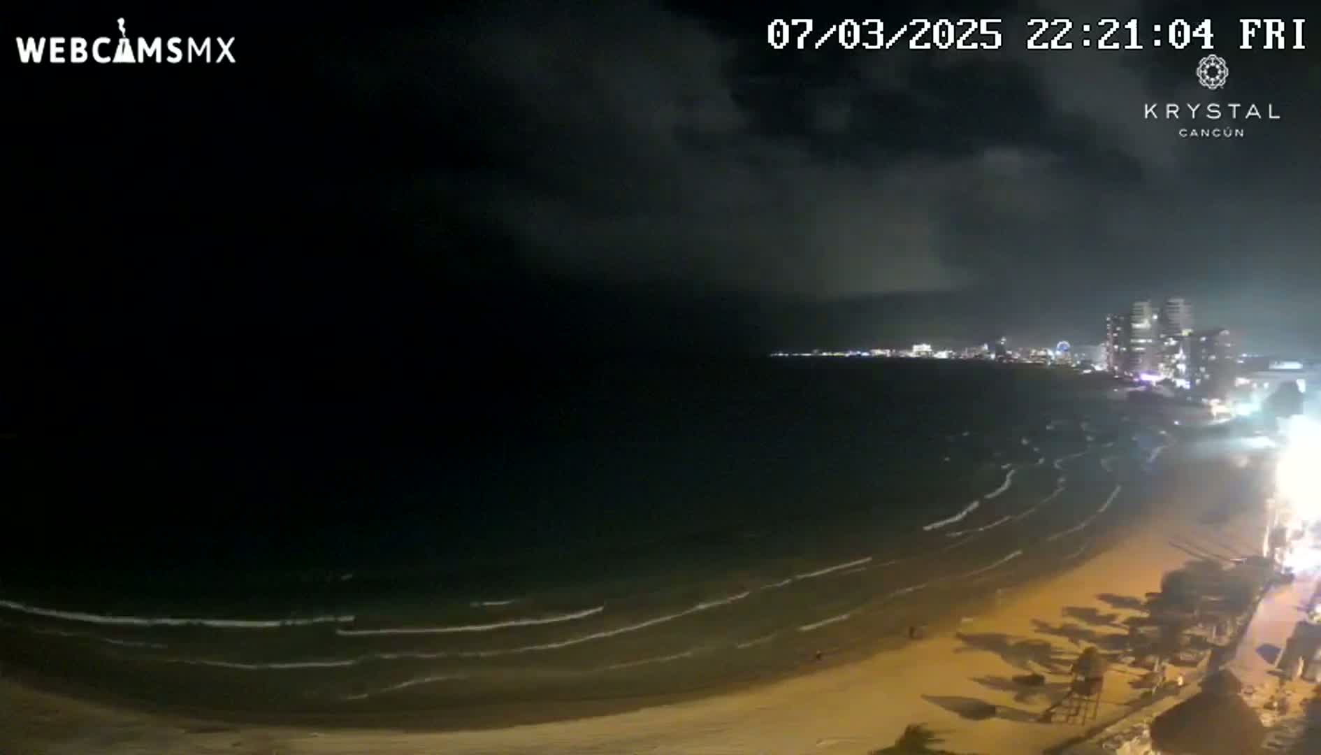 A nighttime beach scene shows gently lapping waves on a sandy shore, under a mostly cloudy sky, with city lights visible in the distance.