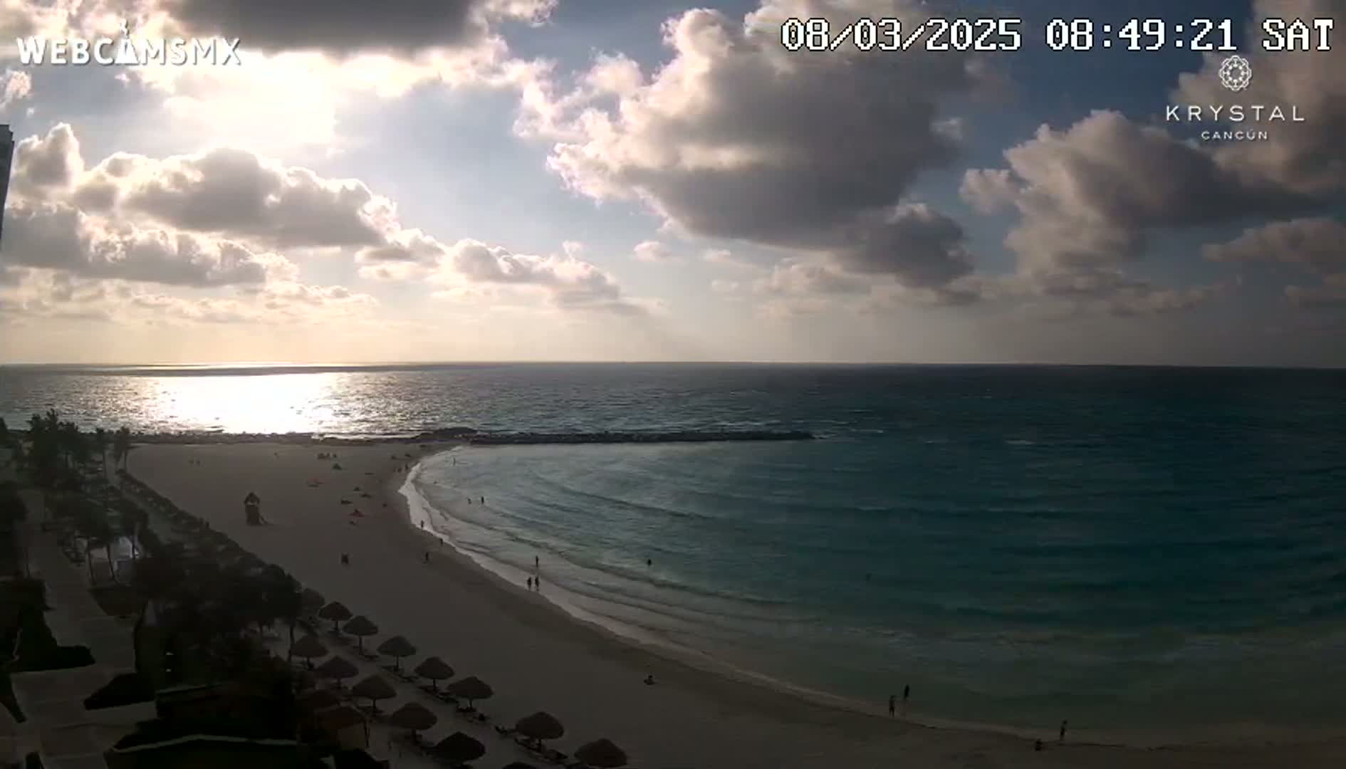 A partly cloudy sunrise over a calm ocean, illuminating a sandy beach with some people and beach umbrellas.