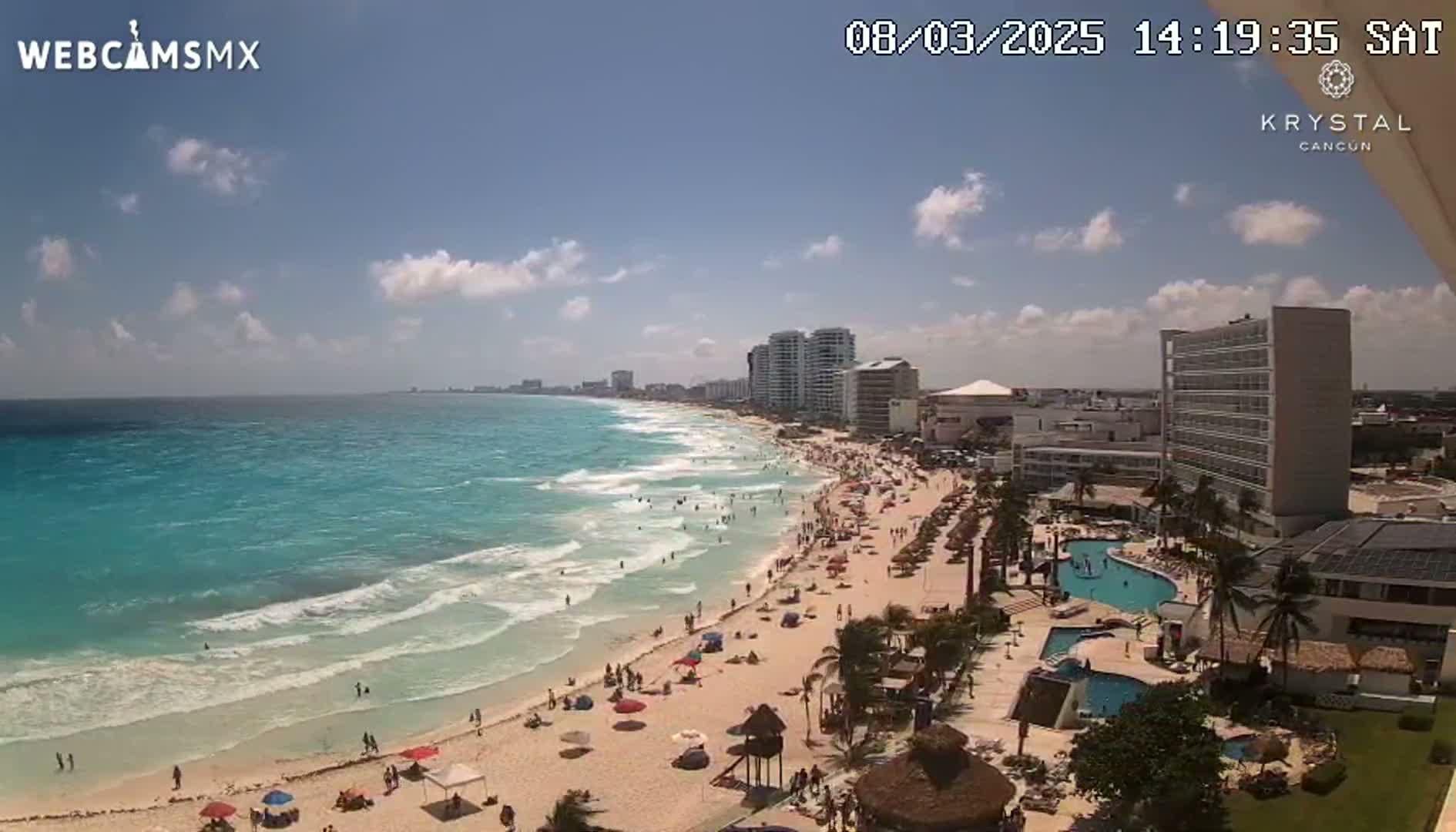 A sunny day shows a crowded beach with turquoise water and waves, hotels, and palm trees.