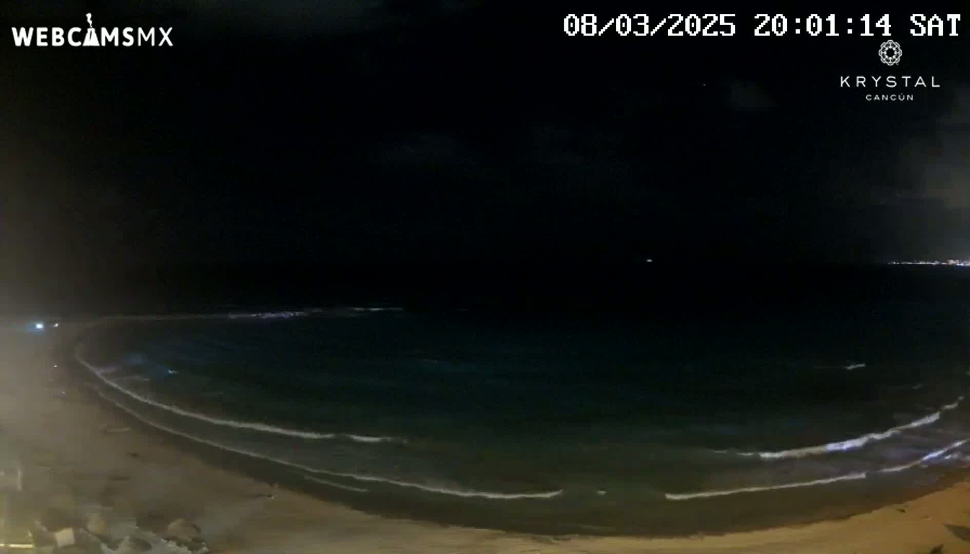 A dark beach at night with gently rolling waves under a mostly clear sky.