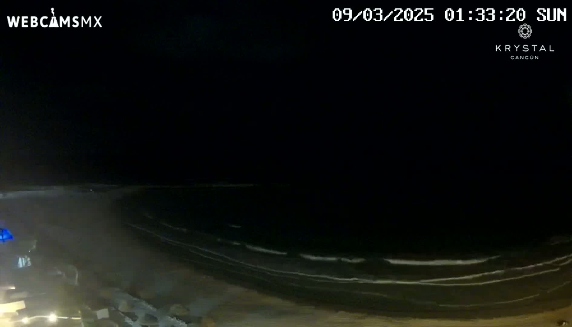 A nighttime, high-angle view shows dark ocean waves lapping a sandy beach.