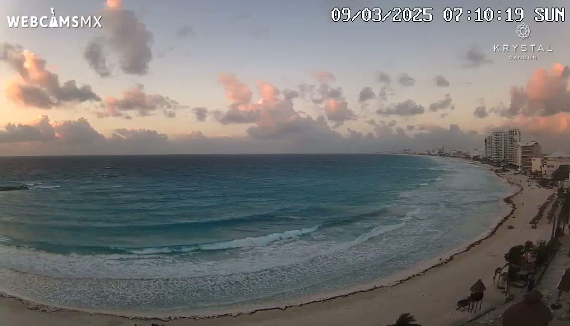 A sandy beach with gentle waves and a partly cloudy sunset overlooks a coastline with buildings in the distance.