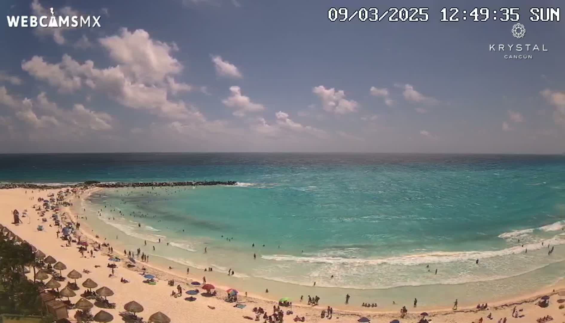 A sunny day at a beach shows a calm, turquoise ocean lapping a sandy shore crowded with people under beach umbrellas.