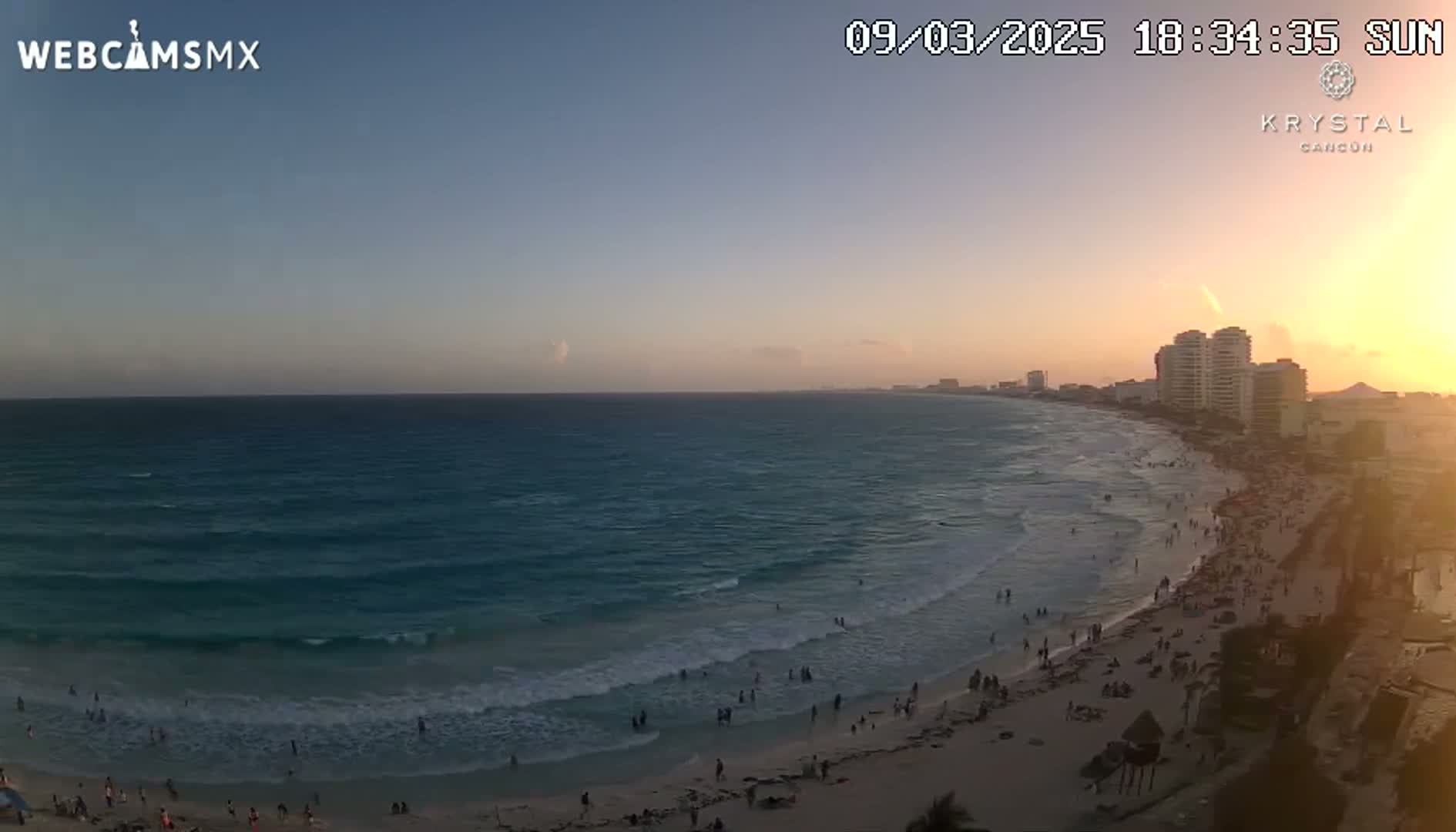 A beach with many people on it during sunset, with calm waves lapping the shore.