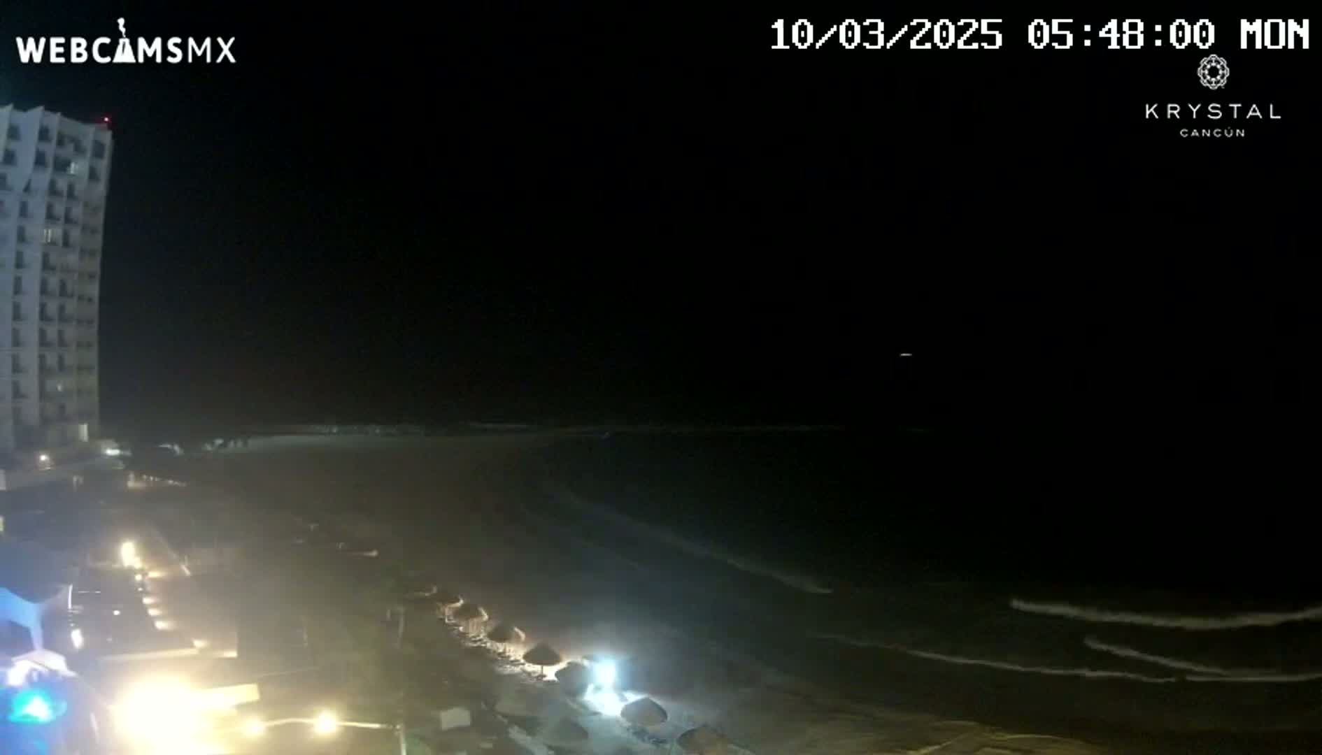 A nighttime view of a beach with gentle waves, a tall building, and some dimly lit beach umbrellas.