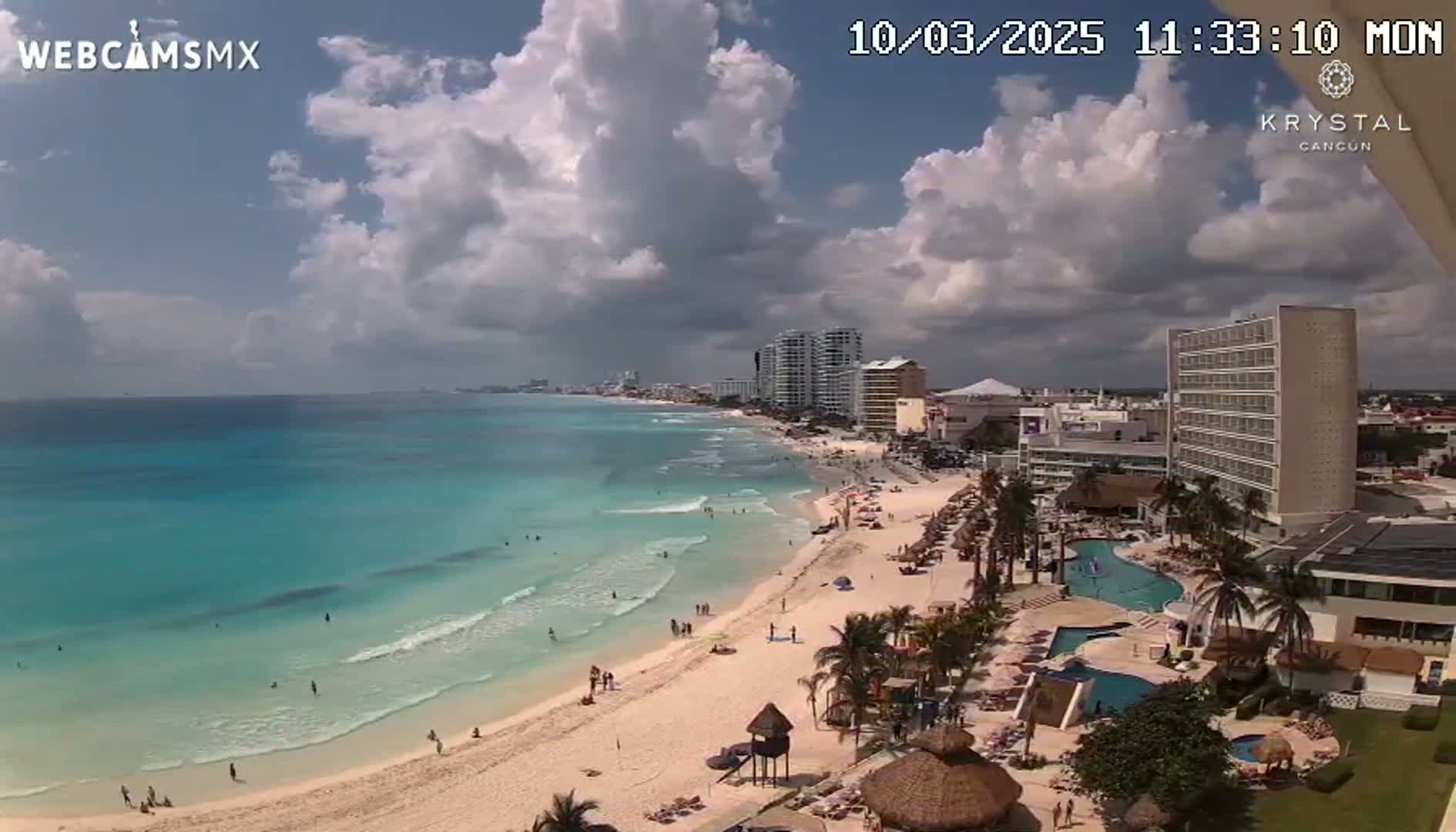 A partly cloudy sky overlooks a beach with turquoise water, several hotels, and palm trees.
