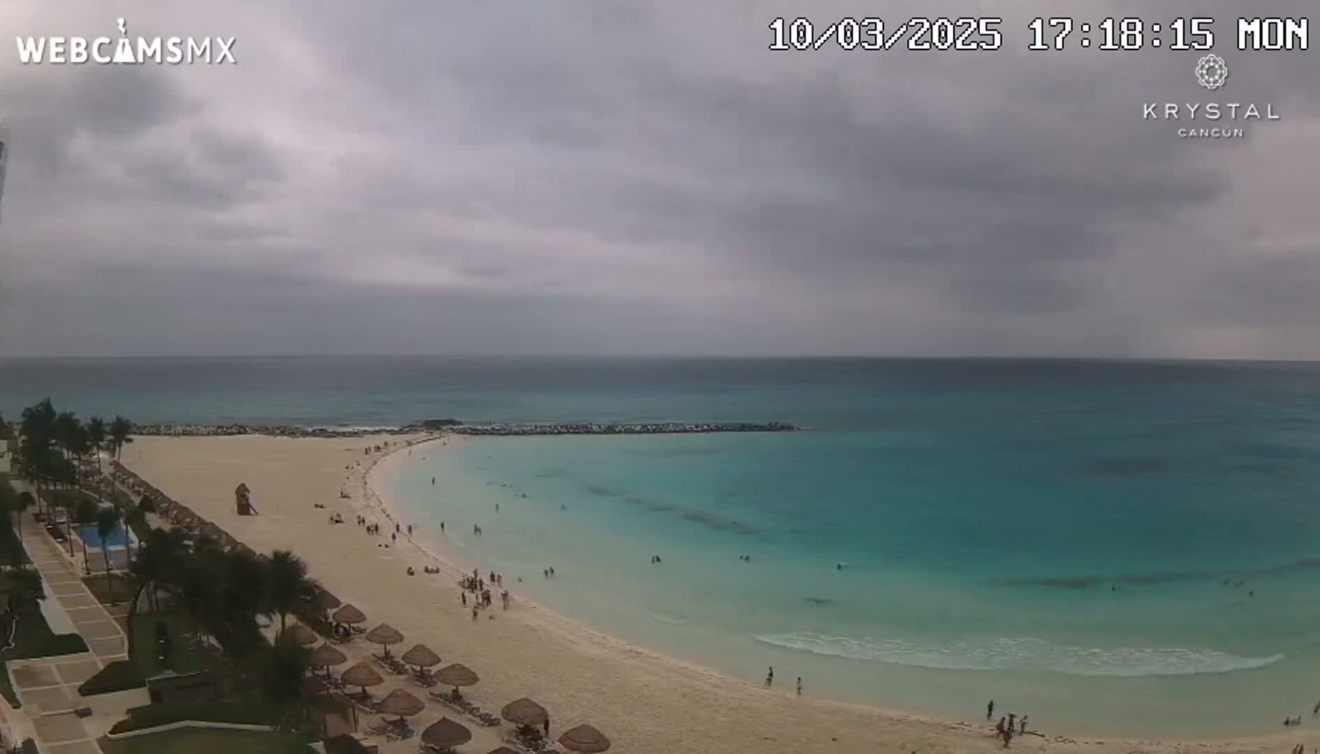 A mostly cloudy sky hangs over a sandy beach with turquoise water, where people are swimming and sunbathing.