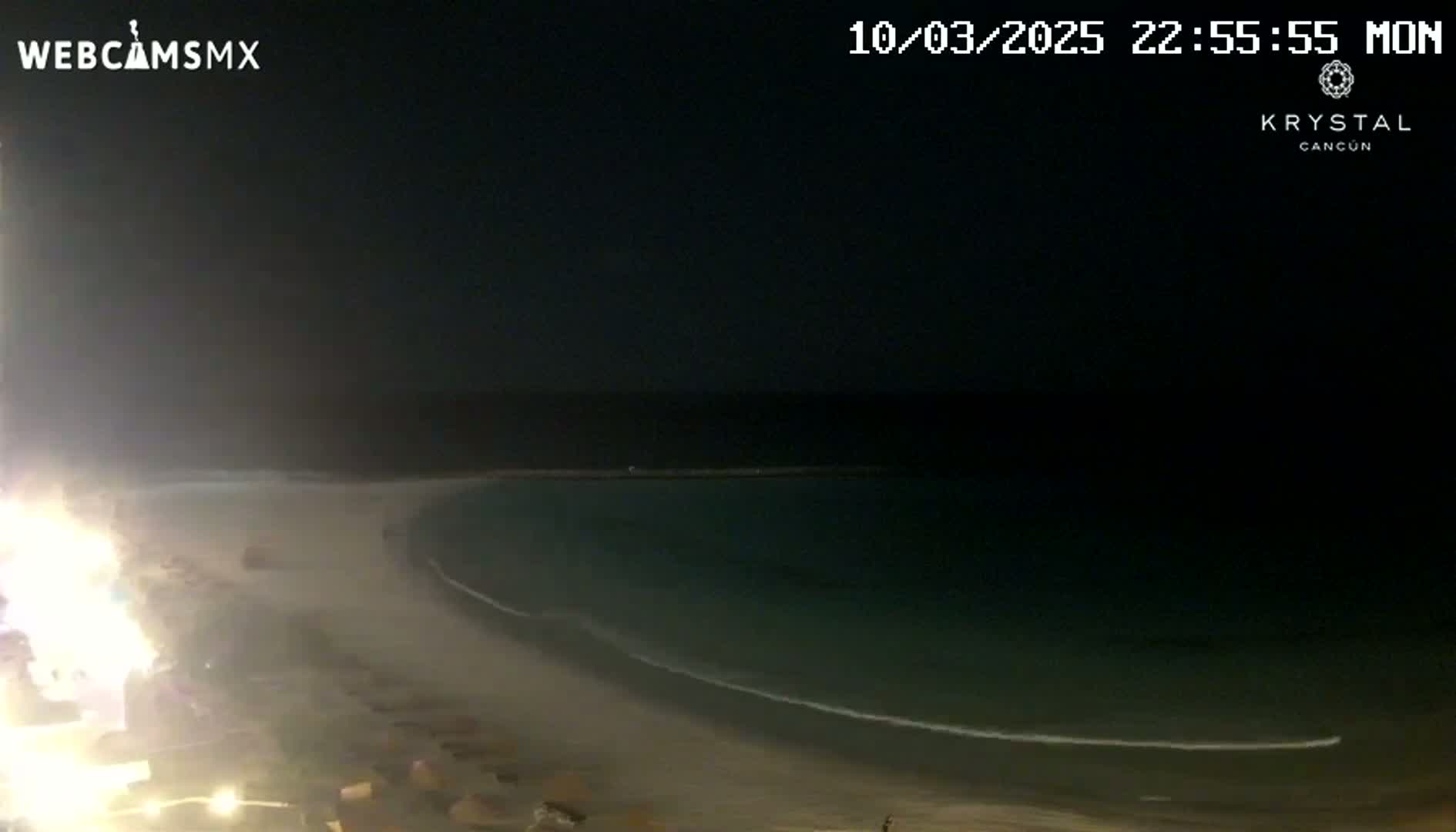A nighttime beach scene shows a calm, dark-blue ocean meeting a light-colored sandy shore under a clear, dark sky.