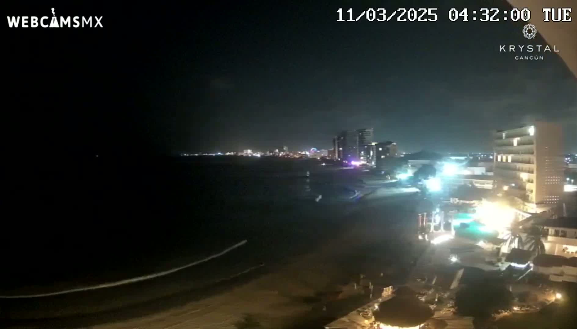 A nighttime view of a beach with a city skyline in the distance, lit by artificial lights, with gently lapping waves on the shore.