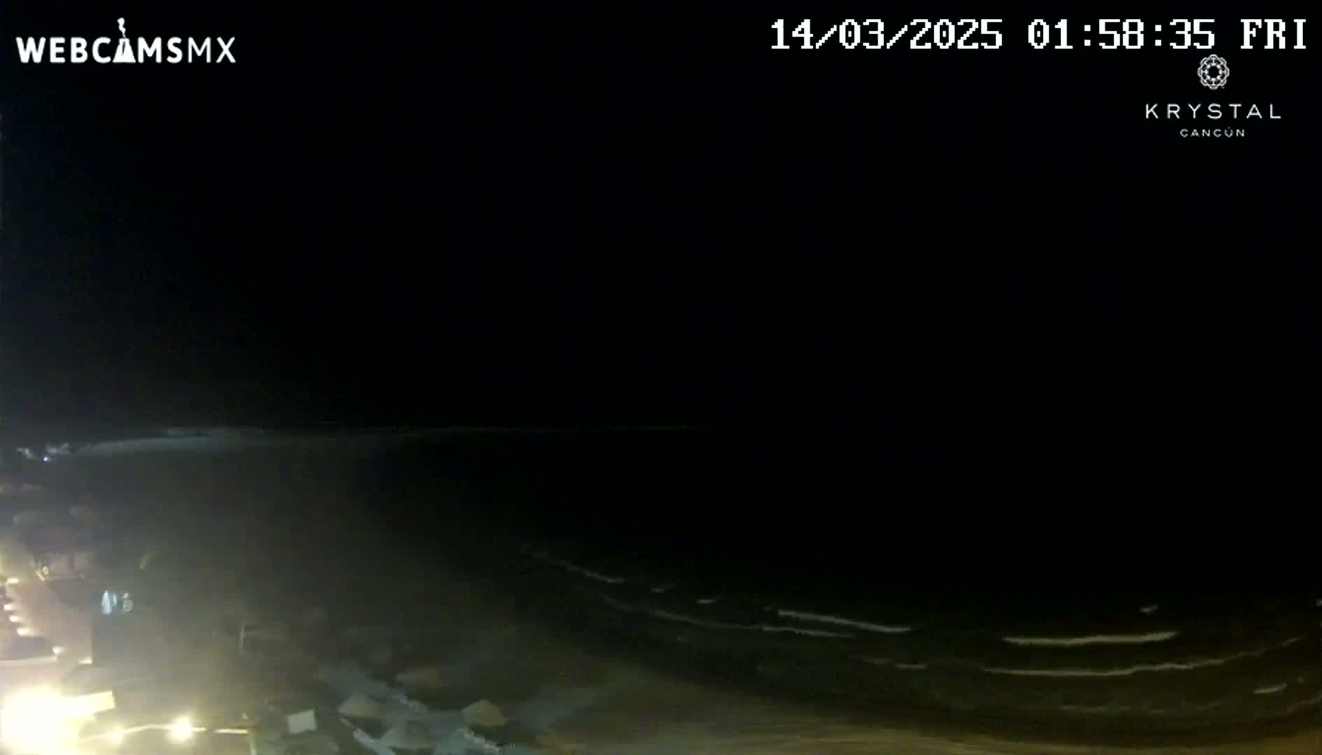 A nighttime, seaside view shows dark water with small waves lapping a dimly lit shore.