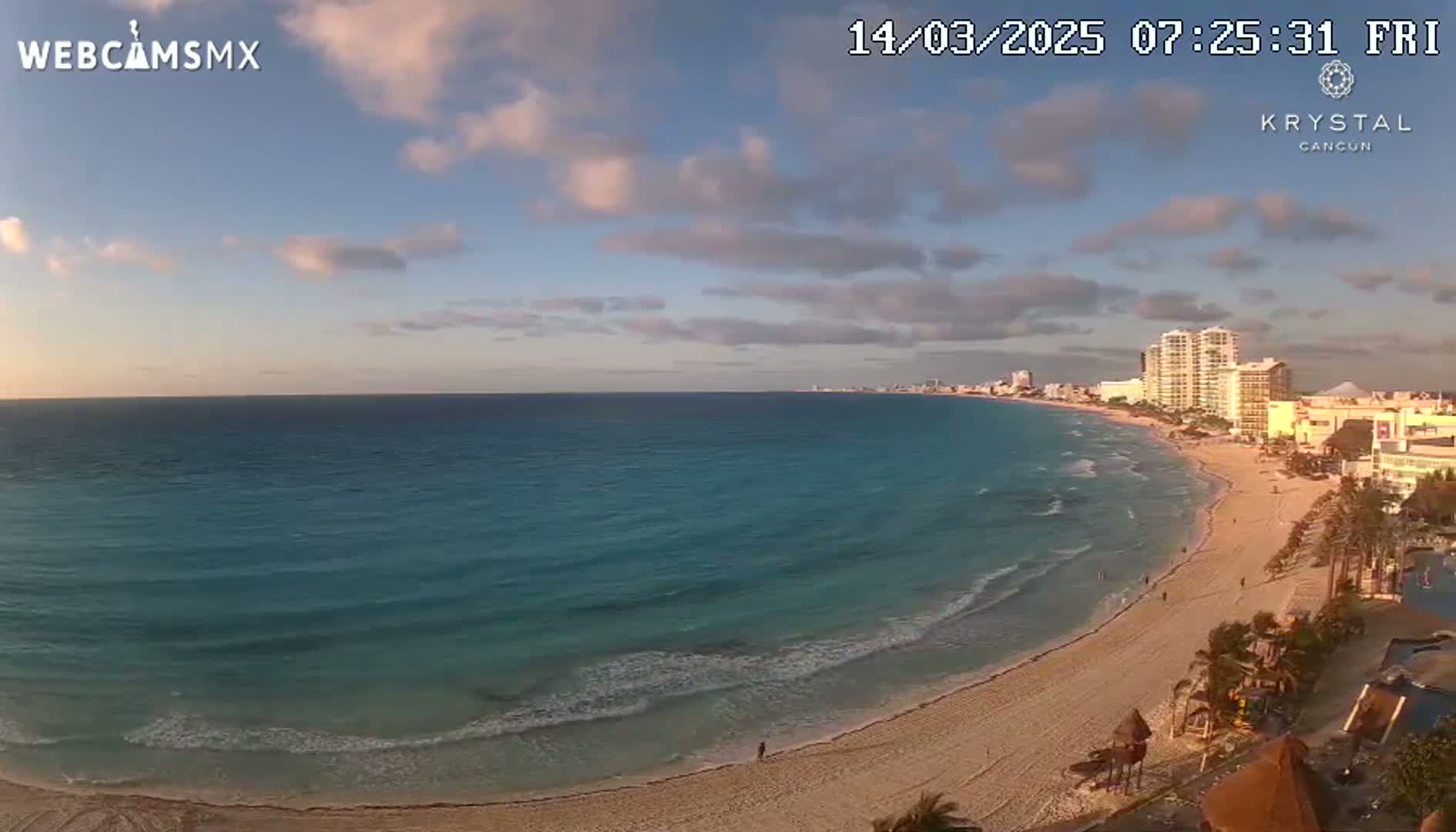 A mostly clear sky with a few clouds overlooks a long sandy beach and calm turquoise ocean, with hotels and buildings visible in the distance.