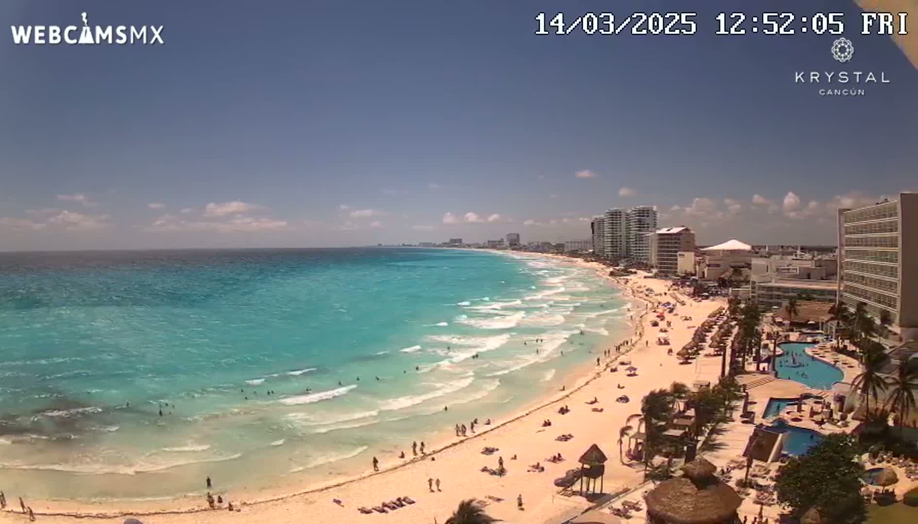 A sunny day on a white sand beach with turquoise water shows many people swimming and sunbathing, with hotels and buildings lining the shore.