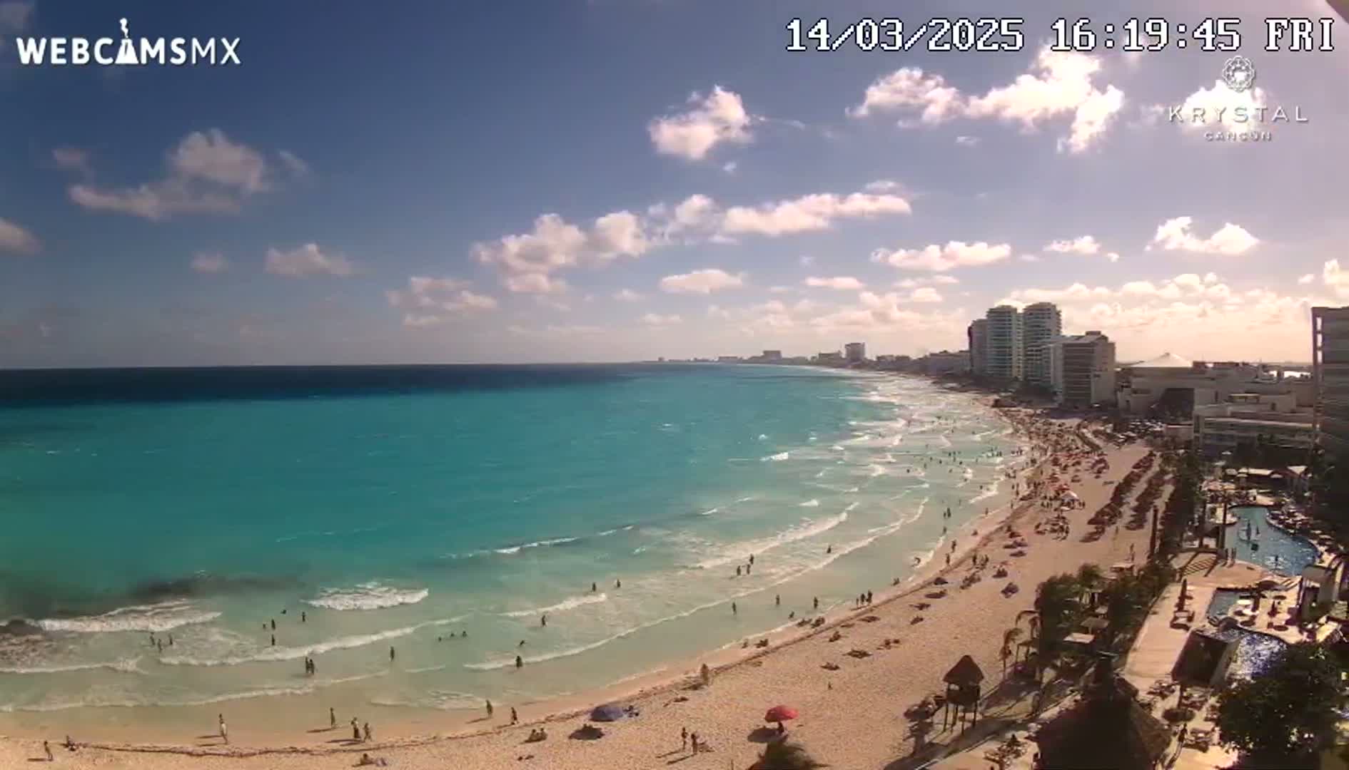 A sunny day shows a beach with turquoise water, waves lapping the shore, and many people enjoying the sand and surf, with a city skyline visible in the background.