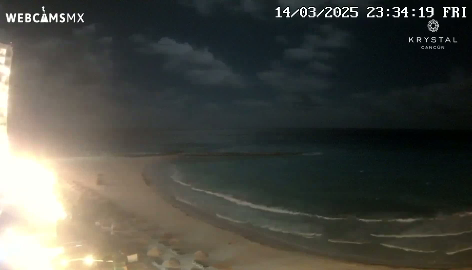 A nighttime beach scene shows a sandy shore with gentle waves lapping the coastline under a partly cloudy sky.