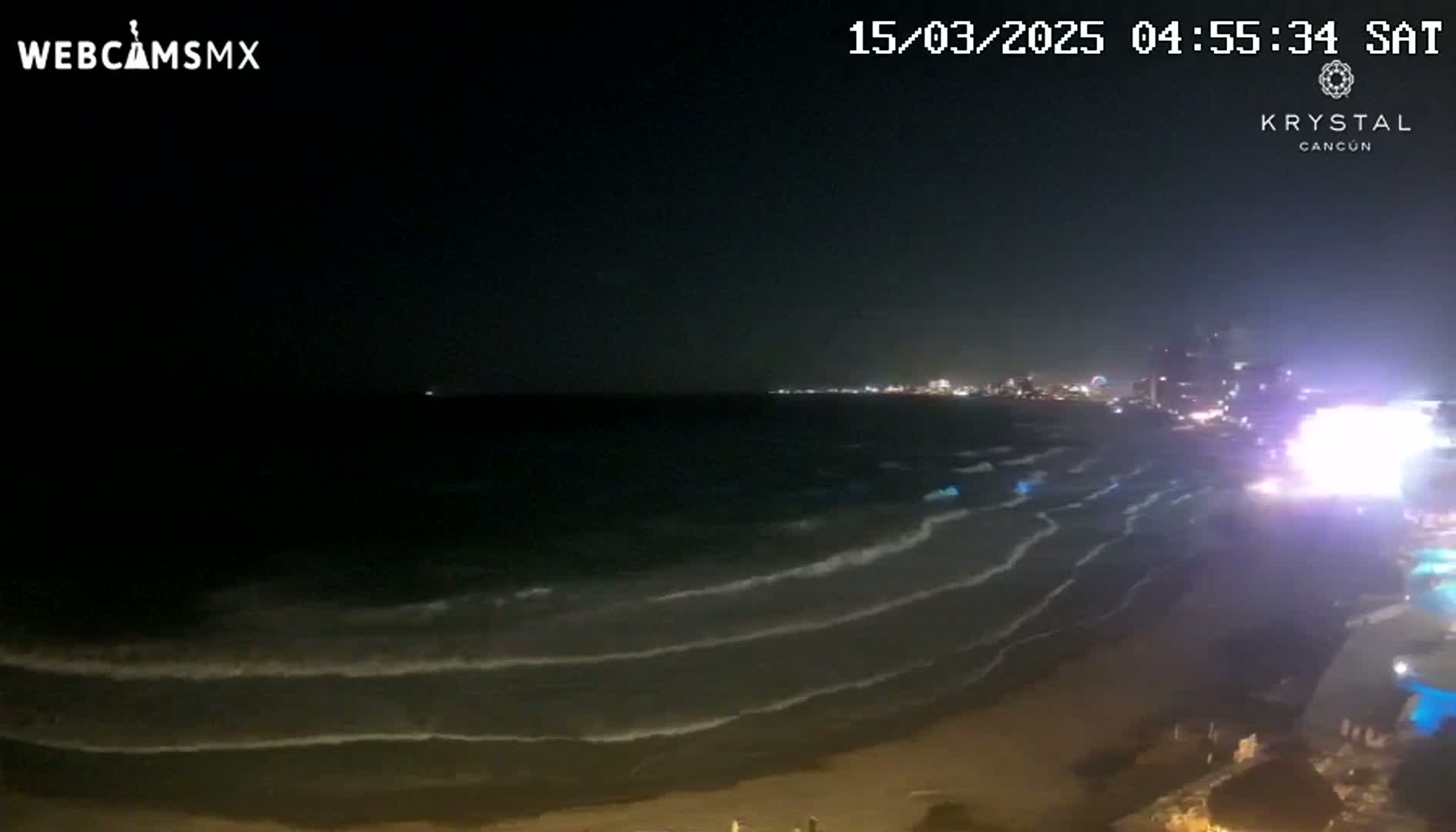 A nighttime beach scene shows gently rolling waves lapping a sandy shore under a dark sky, with city lights visible in the distance.