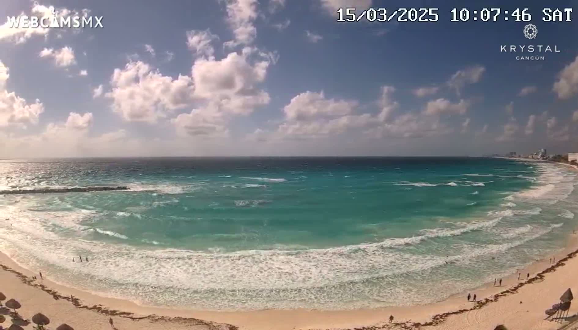 A mostly sunny day shows turquoise water with moderate waves lapping a sandy beach, with a few people visible on the shore.