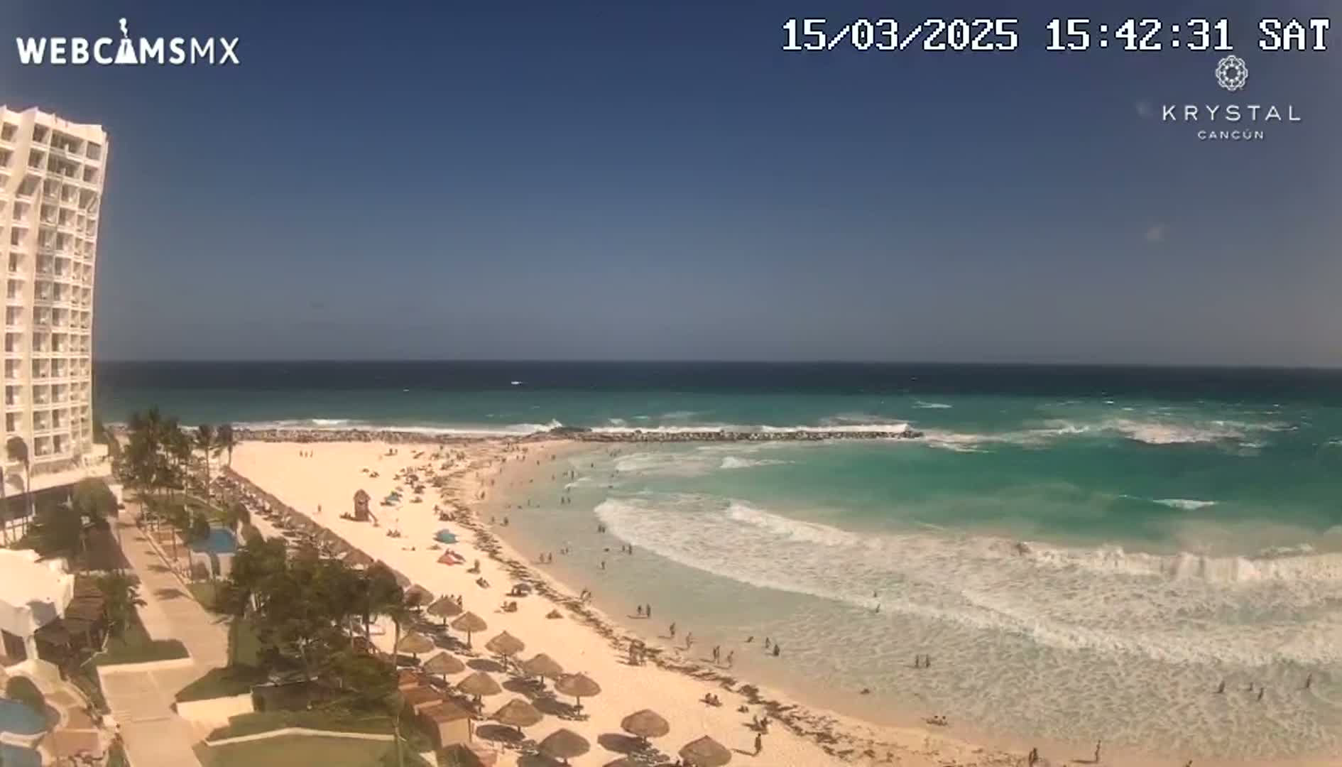 A high-rise hotel overlooks a sunny beach with turquoise water and moderate waves under a clear blue sky.