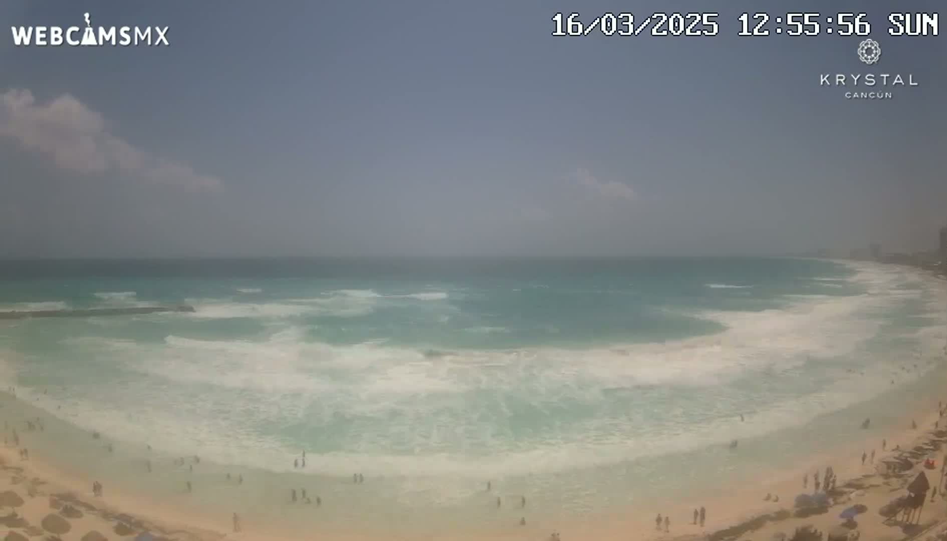 A partly cloudy day shows a long expanse of light teal ocean waves breaking on a sandy beach with a few people scattered along the shore.