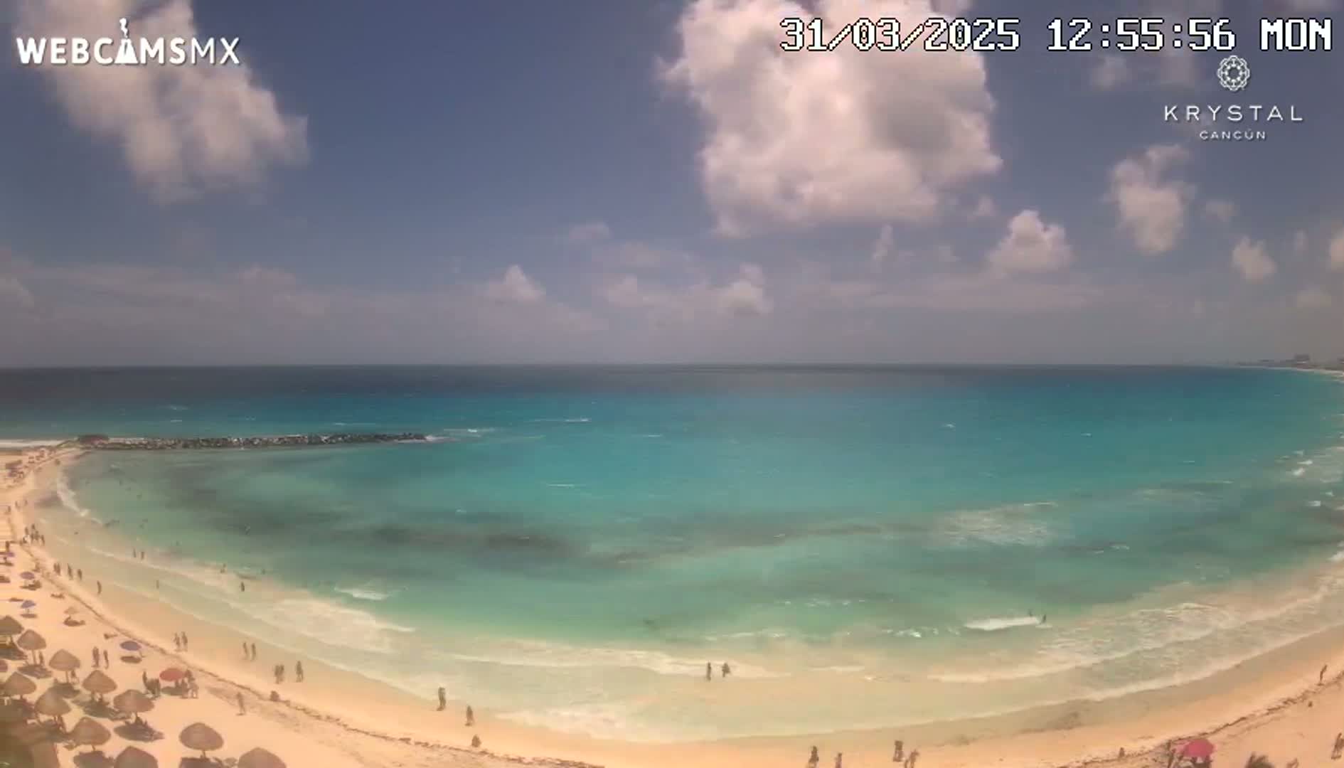 A partly cloudy day shows a beach with turquoise water and light sand, featuring people swimming and relaxing under beach umbrellas.