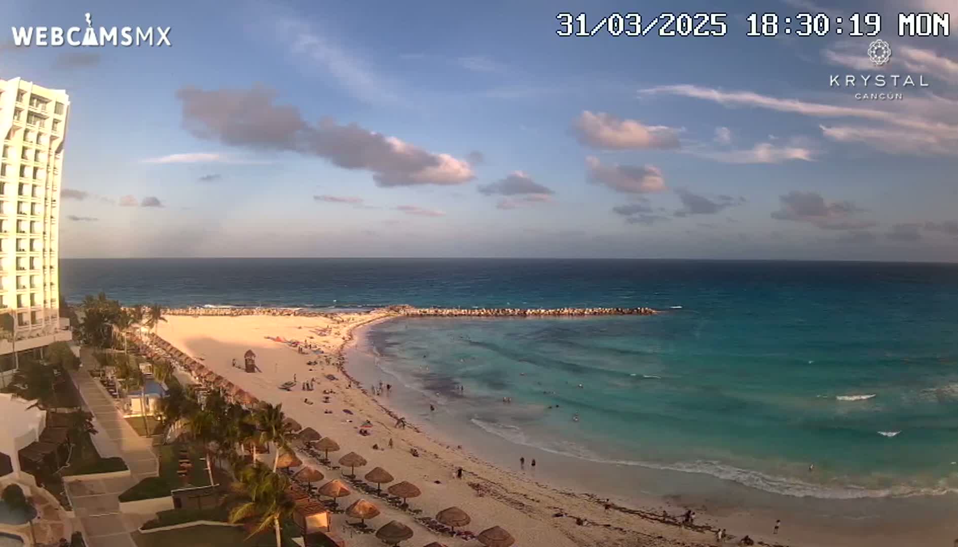 A partly cloudy evening scene shows a beach with people, palm trees, and beach umbrellas next to a tall hotel overlooking a calm turquoise ocean.