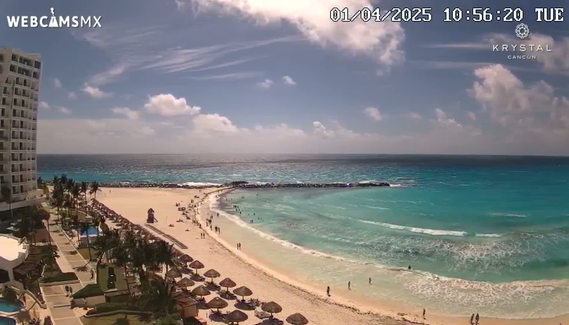 A partly cloudy day shows a beach with turquoise water and a resort with palm trees under a mostly sunny sky.