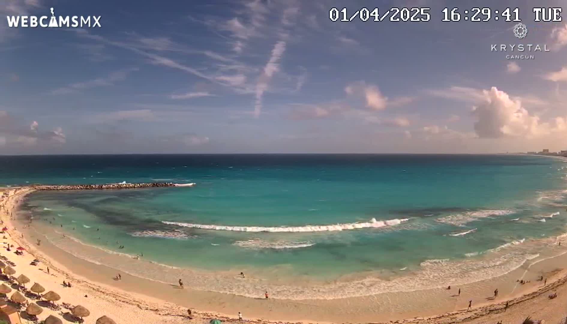 A mostly sunny day shows a beach with turquoise water and small waves lapping a sandy shore where people are swimming and relaxing under beach umbrellas.