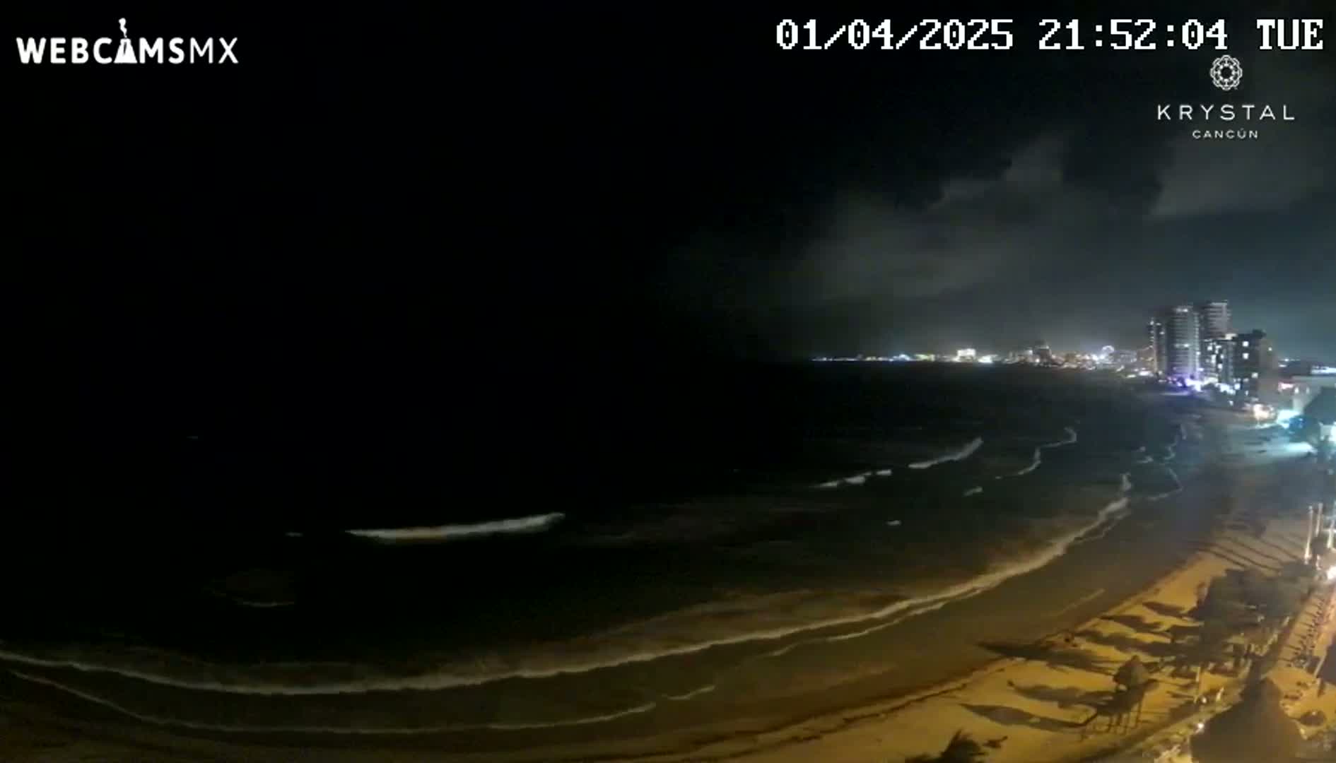 A nighttime beach scene shows dark ocean waves lapping a sandy shore under a partly cloudy sky, with city lights visible in the distance.