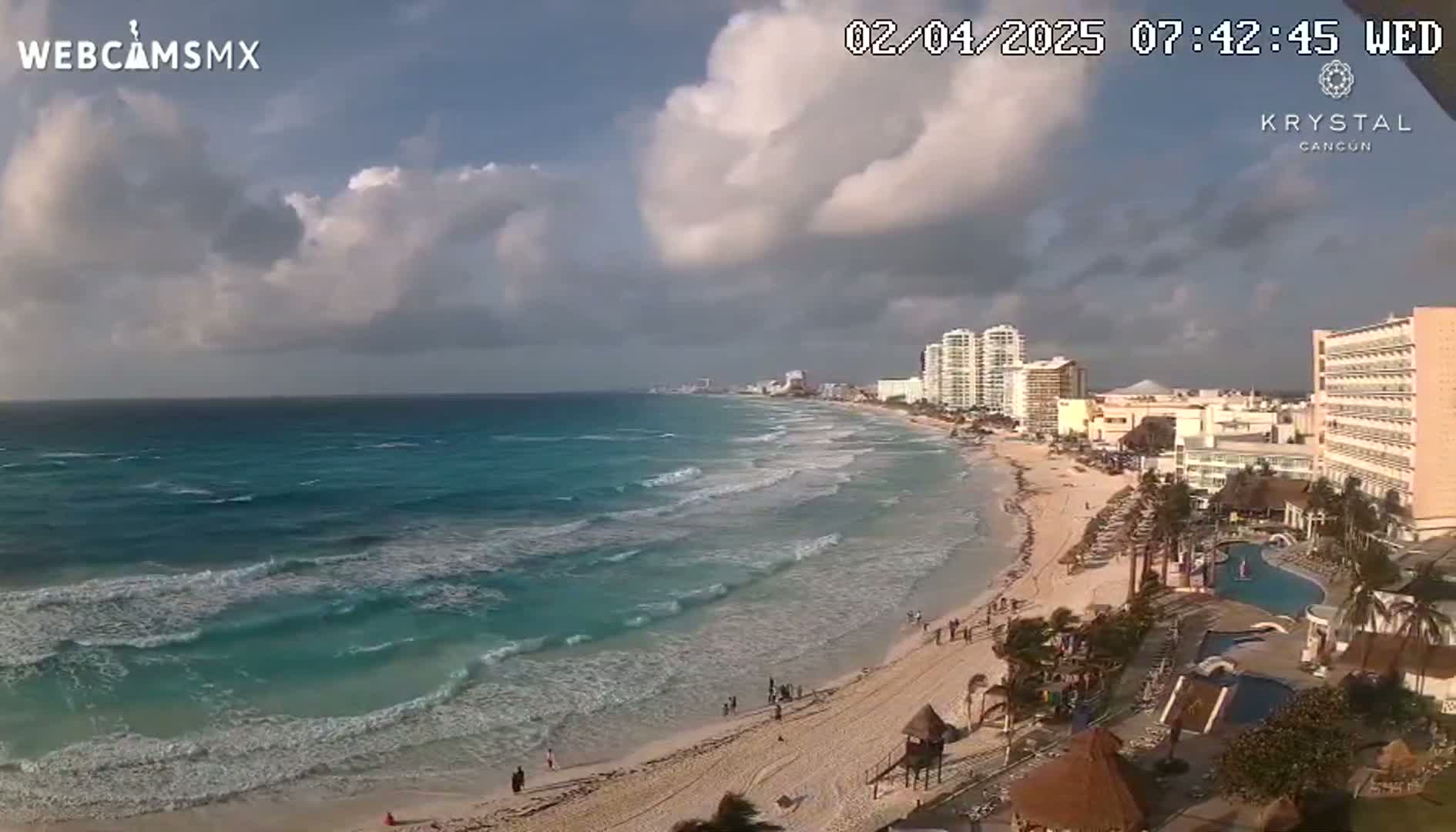 A partly cloudy day shows turquoise ocean waves lapping a sandy beach lined with hotels and resorts.