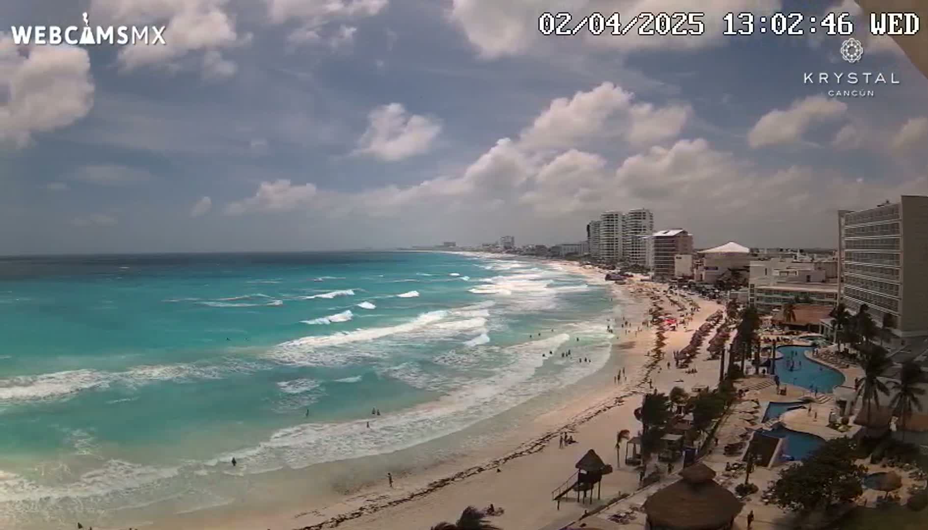 A partly cloudy day shows a beach with turquoise water and waves, along with a shoreline dotted with people and buildings.