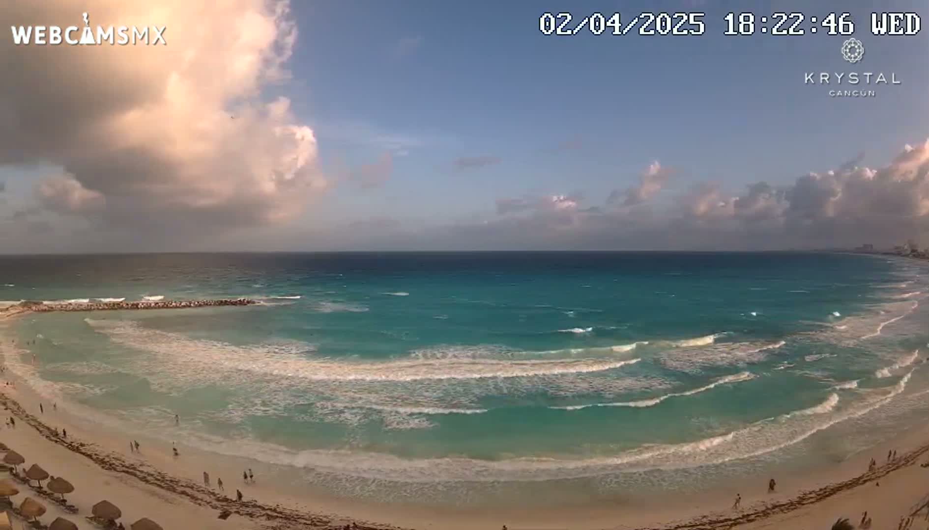 A partly cloudy sunset over a sandy beach with turquoise water and gentle waves.