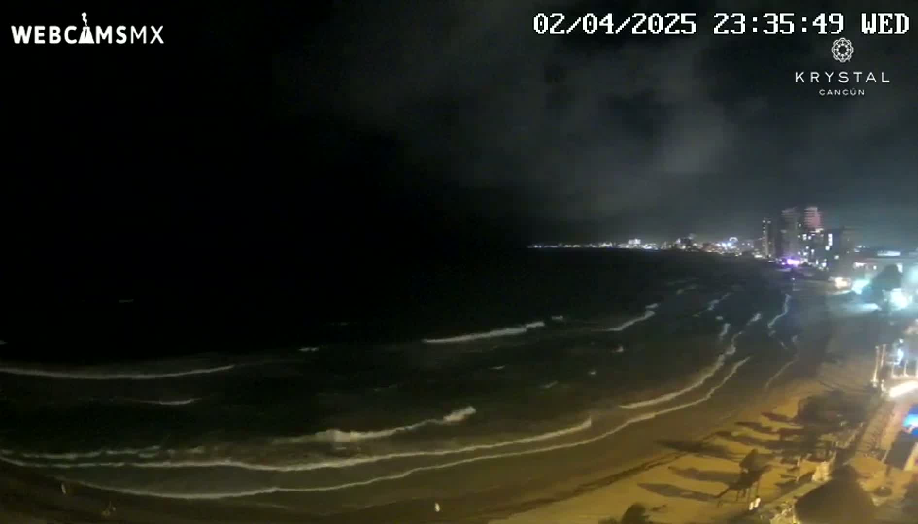 A nighttime, seaside view shows gently rolling waves lapping a dark sandy beach, with a city skyline illuminated in the distance under a partly cloudy sky.