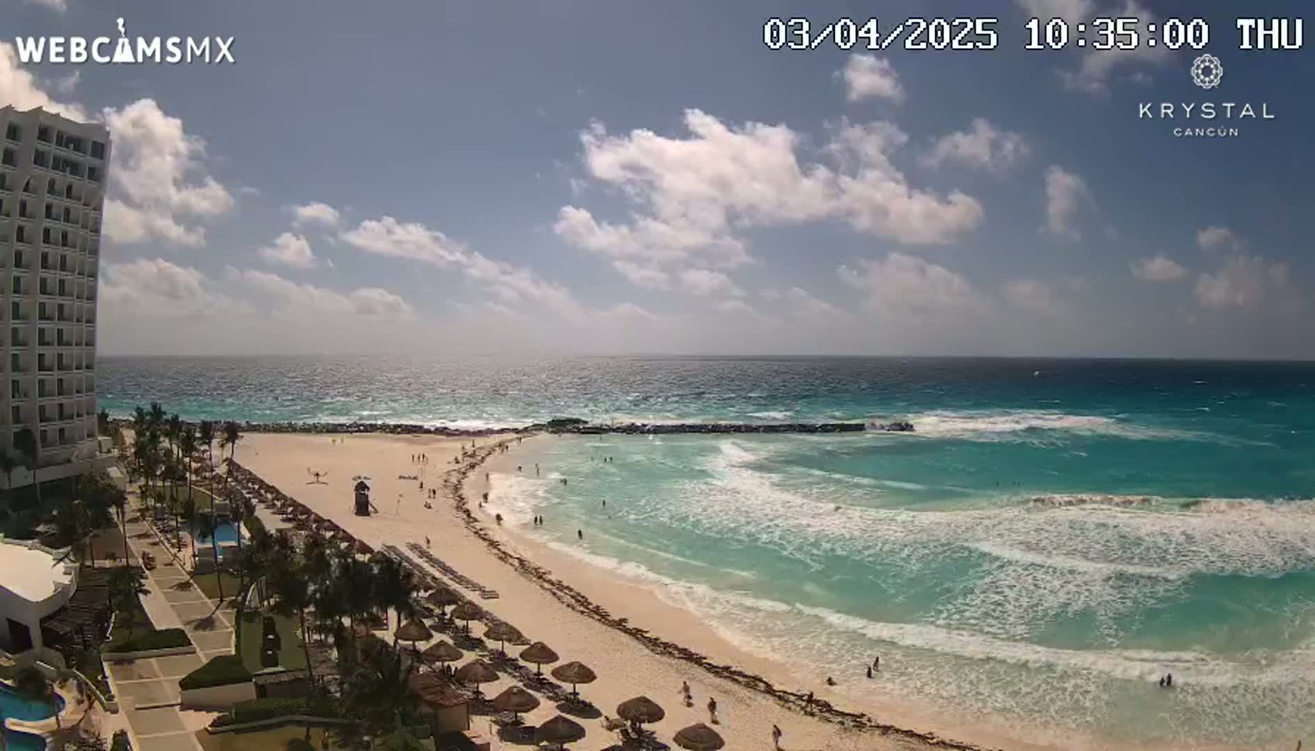 A mostly sunny day shows a beach with turquoise water, waves breaking near the shore, a sandy beach with some people on it, and a tall hotel in the background.