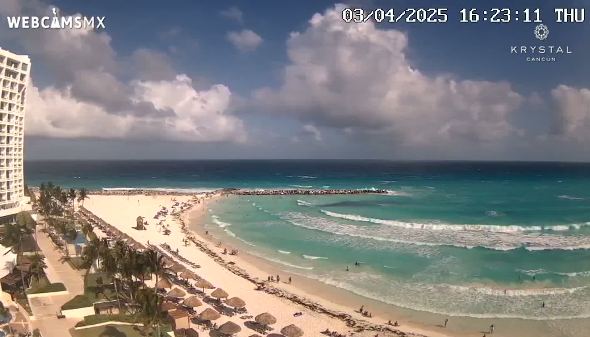 A partly cloudy day reveals a beachfront resort with a sandy beach and turquoise water featuring gentle waves.