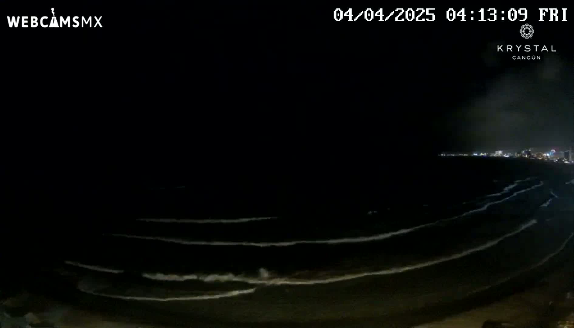 At night, gently rolling waves lap a dark shoreline under a clear, dark sky with a distant city skyline faintly visible.