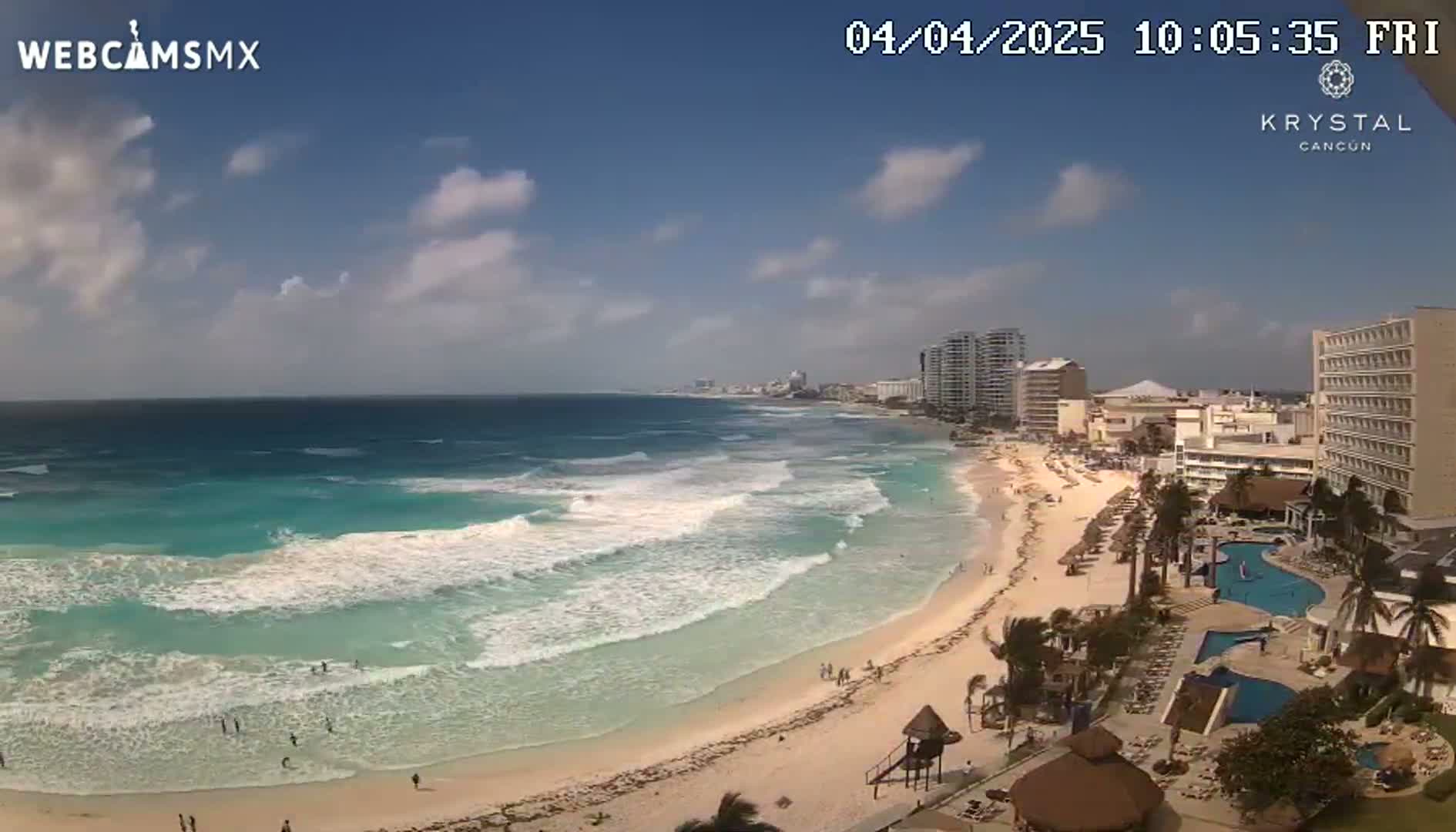 A partly cloudy day shows turquoise ocean waves breaking on a sandy beach, with hotels and a swimming pool visible along the shoreline.