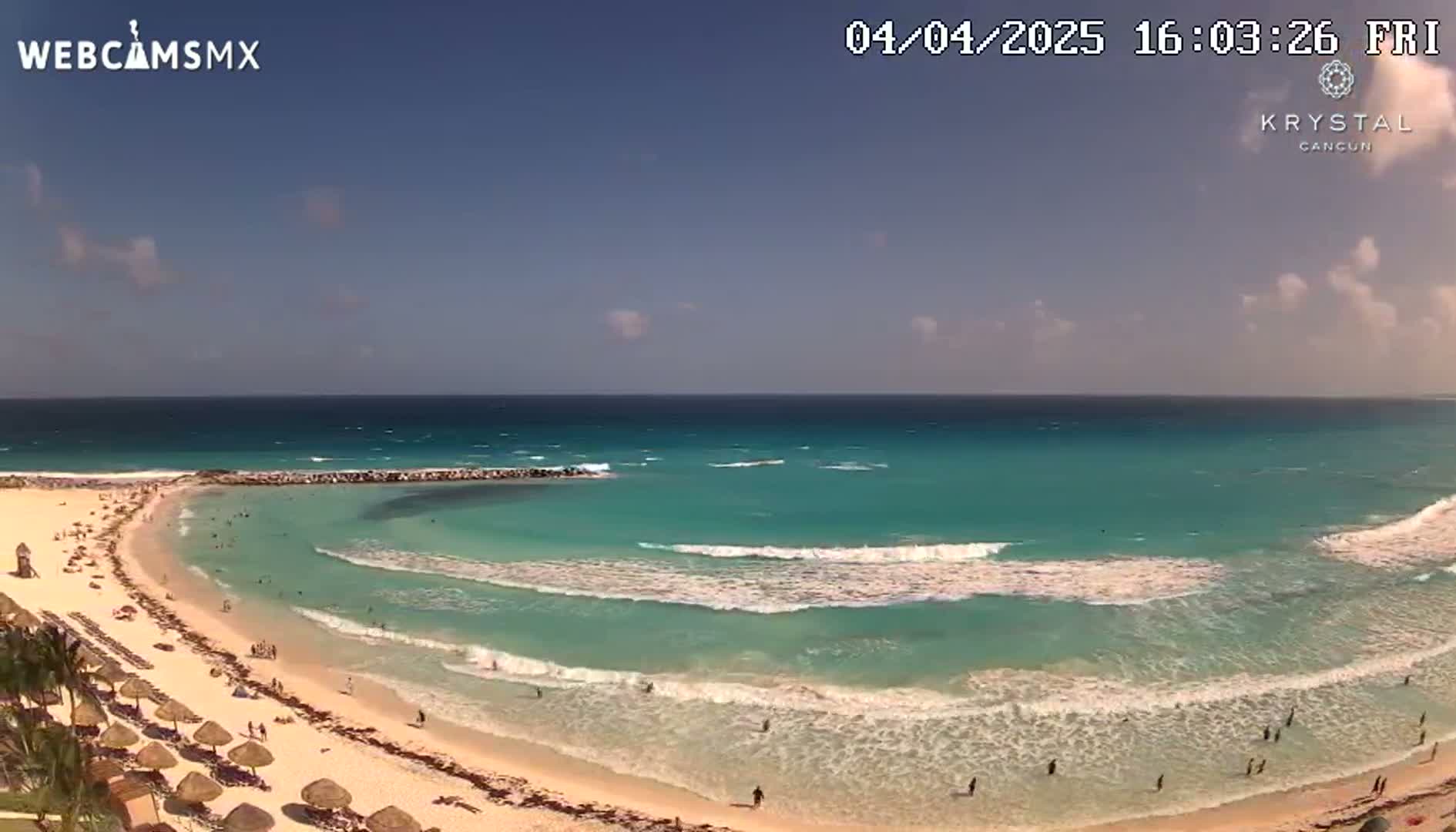 A sunny day shows a sandy beach with turquoise water and small waves, where people are swimming and relaxing under umbrellas.