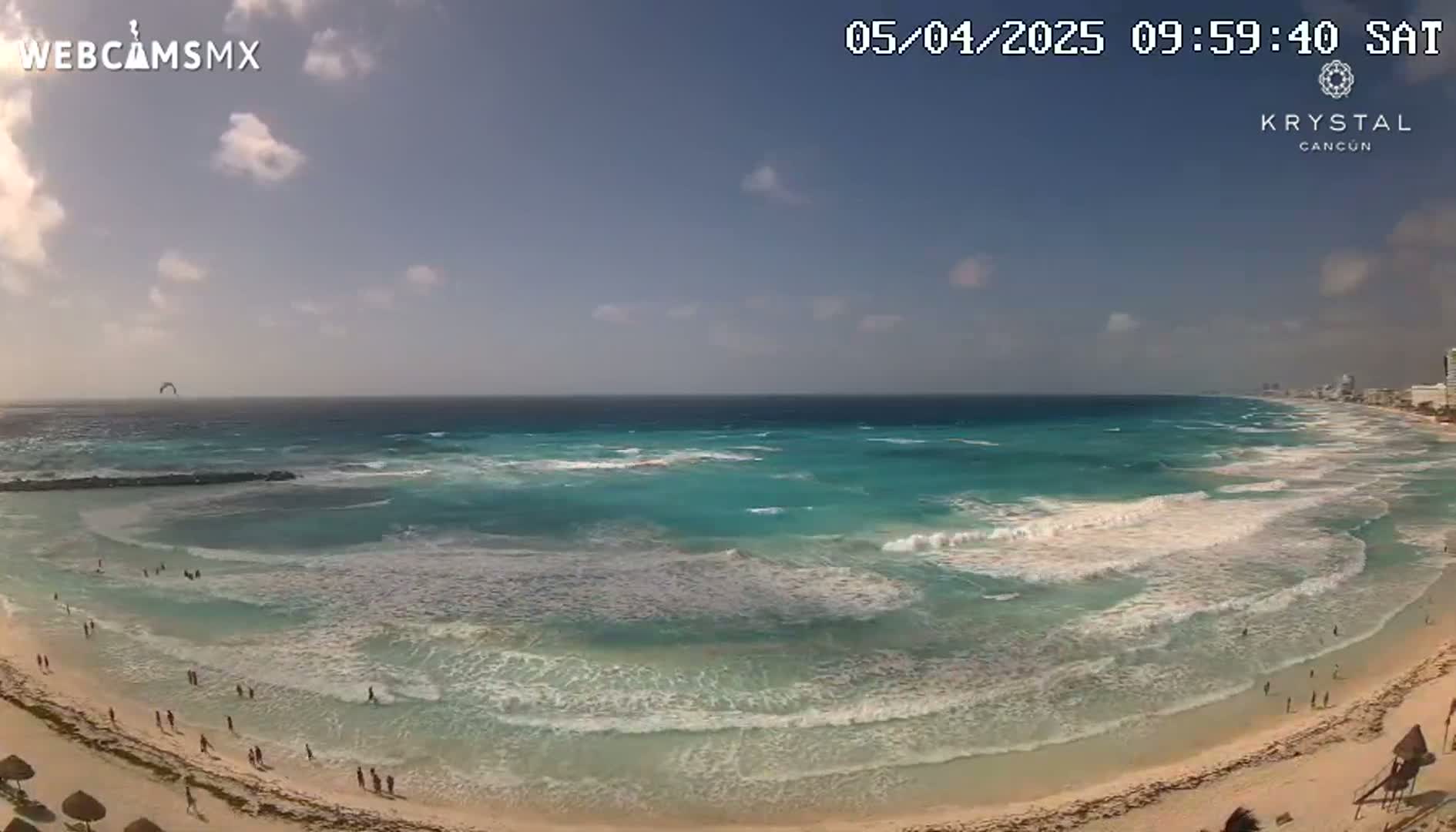 A mostly sunny day shows a wide, sandy beach with turquoise water lapping gently at the shore, featuring people enjoying the beach and a kitesurfer in the distance.