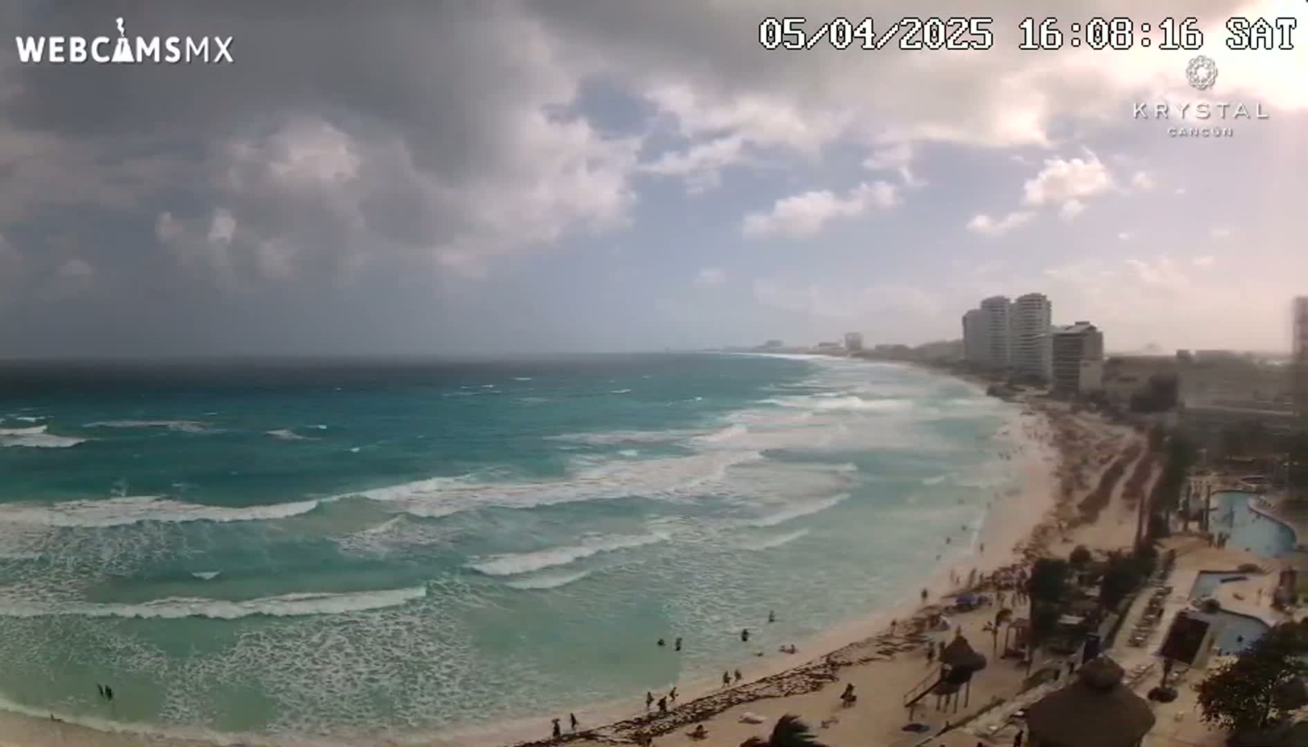 A beach with turquoise water and moderate waves under a partly cloudy sky is seen from an elevated vantage point, showing hotels and people along the shore.
