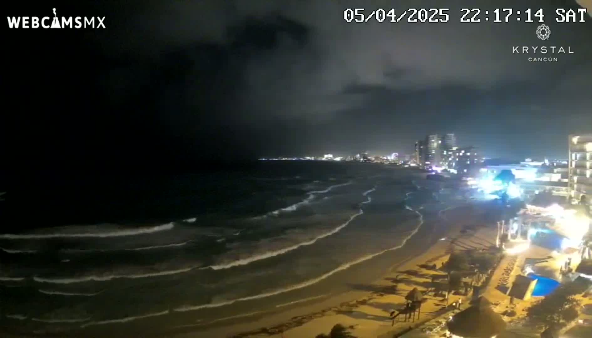 A nighttime beach scene shows moderately sized waves lapping the shore under a cloudy sky.