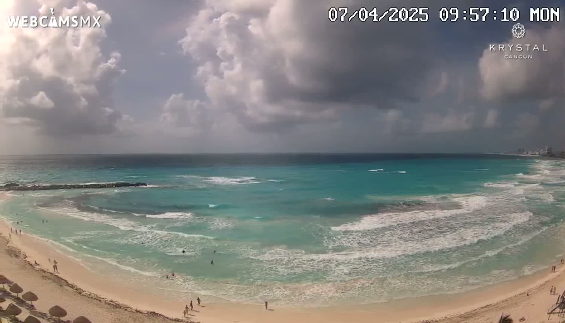 A partly cloudy day shows a sandy beach with turquoise water and small waves lapping the shore, with a few people visible on the sand and in the ocean.