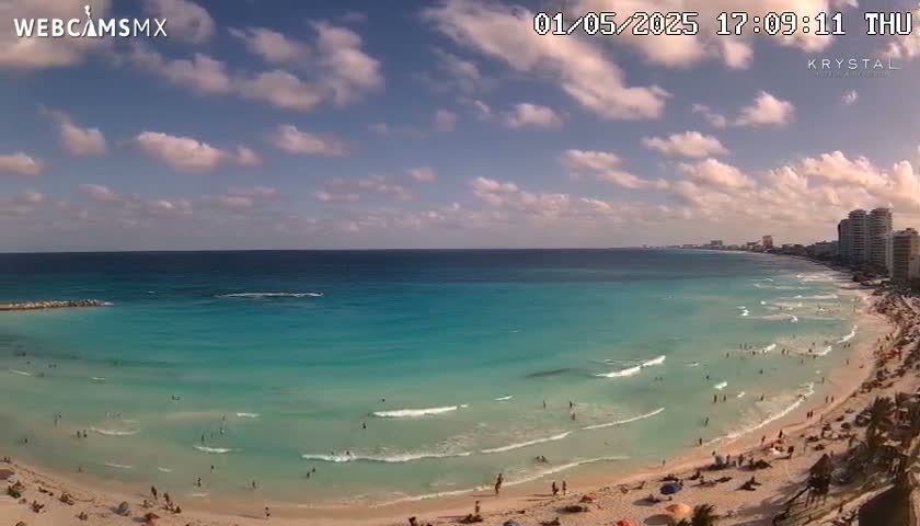A partly cloudy day shows a long sandy beach with turquoise water and many people swimming and sunbathing, with buildings visible in the distance.