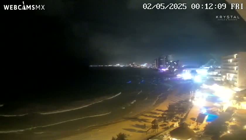 A nighttime beach scene shows dark, gently rolling waves lapping a sandy shore, with illuminated buildings and city lights in the distance under a partly cloudy sky.