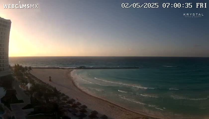 A sunrise casts a warm glow over a sandy beach and calm ocean, with a high-rise building visible to the left.