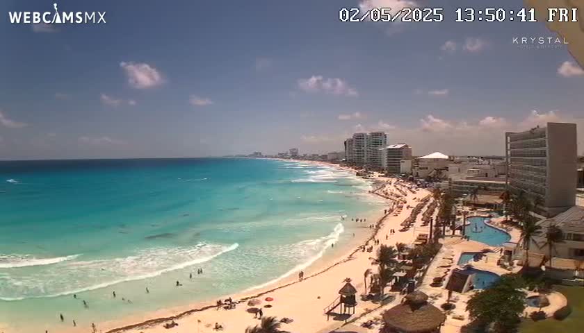A mostly sunny beach scene shows turquoise water lapping a sandy shore lined with beachgoers, hotels, and palm trees.