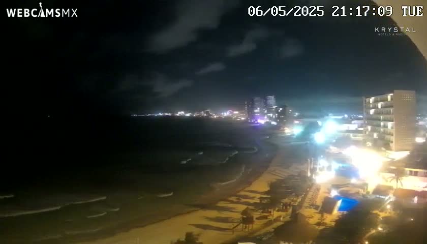 A nighttime view of a beach with gently rolling waves, a brightly lit beachfront, and a city skyline in the distance under a partly cloudy sky.