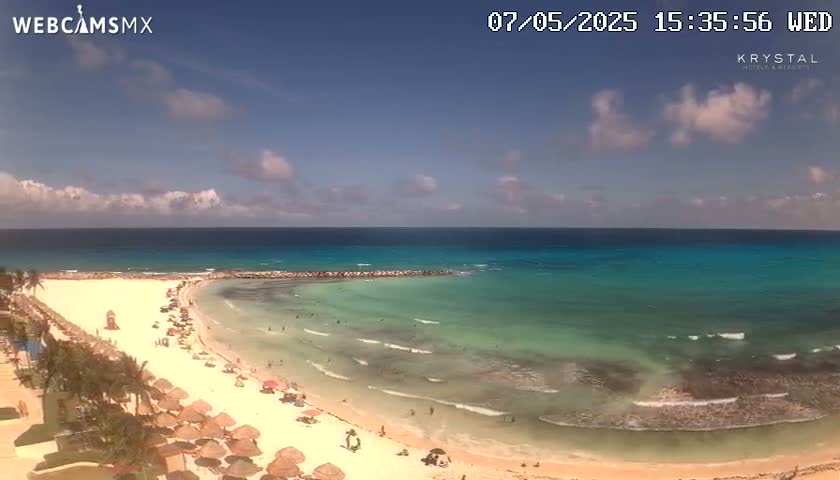 A partly cloudy sunny day shows a beach with turquoise water, small waves, and many people enjoying the sand and sea.